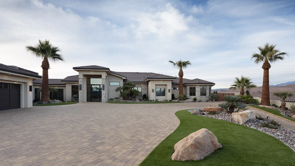Wide paved driveway leading to a home with palm trees and stone accents